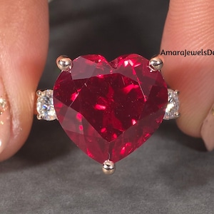 Puede incluir: Un anillo de oro con una gran piedra preciosa roja en forma de corazón. El anillo tiene piedras preciosas más pequeñas y claras a cada lado de la piedra en forma de corazón. El anillo se sostiene sobre un fondo gris.