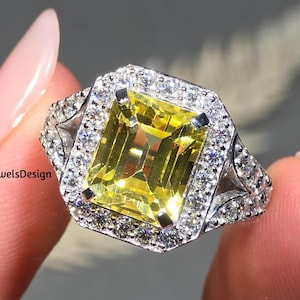 Könnte beinhalten: Ein silberner Ring mit einem großen, rechteckigen gelben Edelstein, umgeben von einem Halo aus kleineren, klaren Edelsteinen. Der Ring hat aufwendige Details am Band und um den Halo. Der Ring wird von einer Hand gehalten.