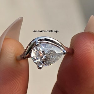 Puede incluir: Un anillo de diamante de forma de pera con una banda de plata. El anillo está engastado en una banda curva y presenta un diamante grande y claro.