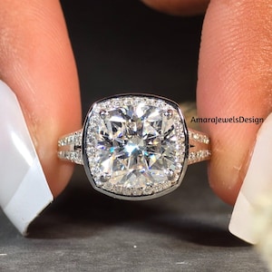 May include: A silver ring with a large square-shaped diamond surrounded by smaller diamonds. The ring has a double band with smaller diamonds on each band. The ring is on a finger with white nail polish.