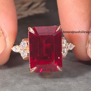 Puede incluir: Un anillo de oro con una gran piedra preciosa roja oscura rectangular como pieza central. El anillo está adornado con piedras preciosas más pequeñas y claras a ambos lados de la piedra principal. El anillo está sostenido por los dedos de una persona.