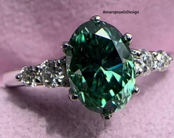 May include: A silver ring with a large oval green gemstone surrounded by smaller white gemstones. The ring is on a pink background. AmarajewelsDesign is written in the top right corner of the image.