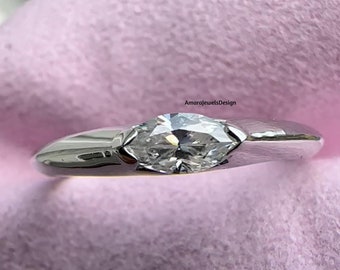 May include: A marquise-cut diamond ring with a polished, silver-colored band. The diamond is set horizontally, and the ring is displayed on a pink surface. The text "AmarajewelsDesign" is visible.