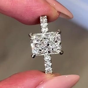 May include: A silver ring featuring a large, square-shaped diamond centre stone. The diamond is surrounded by smaller diamonds that form a halo around the centre stone. The band of the ring is also set with smaller diamonds.