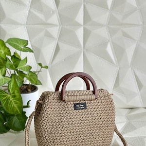 May include: A beige crochet handbag with brown wooden handles and a detachable shoulder strap. The bag features a rectangular shape with a textured stitch pattern and a small black label. The bag is set against a white geometric background.