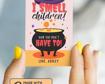 May include: A Halloween-themed card held by a person with yellow nail polish. The card features a purple witch hat, a black cauldron, and the text "I SMELL children! NOW YOU DON'T HAVE TO! LOVE, ASHLEY."