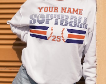 May include: White sweatshirt with a blue and orange softball design. The text "YOUR NAME SOFTBALL" is printed on the front of the sweatshirt, along with a baseball and the number 25.