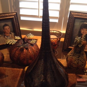May include: A tall, dark brown ceramic vase with a textured surface. The vase is decorated with a subtle metallic sheen. It is displayed on a table with other decorative items, including a pumpkin and a gourd.