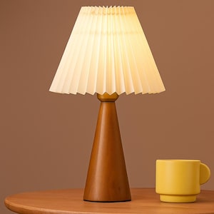 May include: A wooden table lamp with a pleated off-white lampshade. The lamp has a conical wooden base and a gold-colored neck. A yellow mug sits on the table next to the lamp.