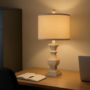 May include: A beige table lamp with a fabric shade and a decorative base. The lamp has a round shade with a light-colored fabric and a decorative base. The lamp is on a wooden desk with a notebook and pen.