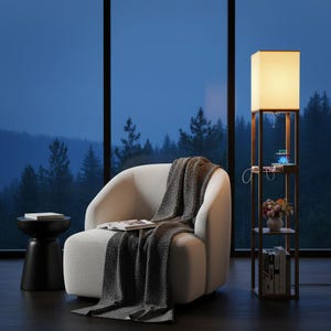 May include: A cozy living room scene featuring a white armchair with a gray blanket and a black side table. A wooden floor lamp with a square lampshade and shelves holds books and decorative items. A large window overlooks a forest.