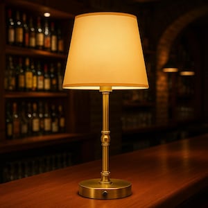 K&ouml;nnte beinhalten: Eine Tischlampe aus Messing mit beigem Lampenschirm. Die Lampe hat einen zylindrischen Sockel und einen schlanken Stiel mit einem dekorativen Detail. Der Lampenschirm hat eine klassische konische Form mit goldenem Rand. Die Lampe steht auf einer Holzoberfl&auml;che.
