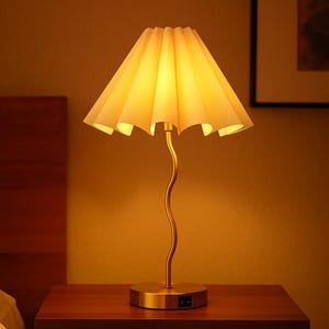 May include: A gold-toned table lamp with a pleated lampshade. The lamp has a curved stem and a round base. The lampshade is a light beige color, and the lamp is turned on, emitting a warm glow. The lamp is on a wooden nightstand.