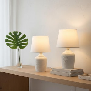 May include: Two white table lamps with textured bases and off-white shades sit on a light wood table. A green monstera leaf in a clear vase is to the left. A small tray and books are also on the table.