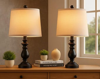 May include: Two black table lamps with white fabric lampshades. The lamps are on a wooden table with a book titled "Interiors Now!" and a small potted plant.