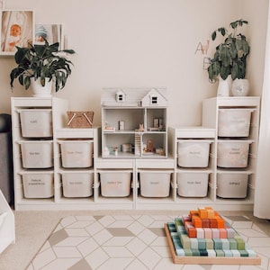 May include: A playroom featuring white storage units, a doll's house, and a wooden block set. The storage units have labelled bins. A geometric patterned rug and framed photos enhance the room's decor. Plants in white pots are placed on top of the storage units.