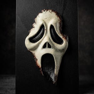 Könnte beinhalten: Skulptur einer Scream-Maske mit einem verwitterten, cremefarbenen Gesicht, schwarzen Augenhöhlen und Mund, montiert auf einem schwarzen rechteckigen Sockel. Die Maske hat ein gealtertes Aussehen mit braunen Akzenten.