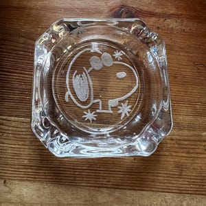 May include: Clear glass ashtray with a Snoopy design etched in the center. The ashtray has a square shape with rounded corners and a scalloped edge. The Snoopy image is white and detailed.