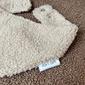 May include: A beige sherpa bib with a textured, fleece-like surface. The bib features two silver-toned metal snap closures at the neck and a small white fabric tag on the side with the text marli carli printed in black lowercase letters.
