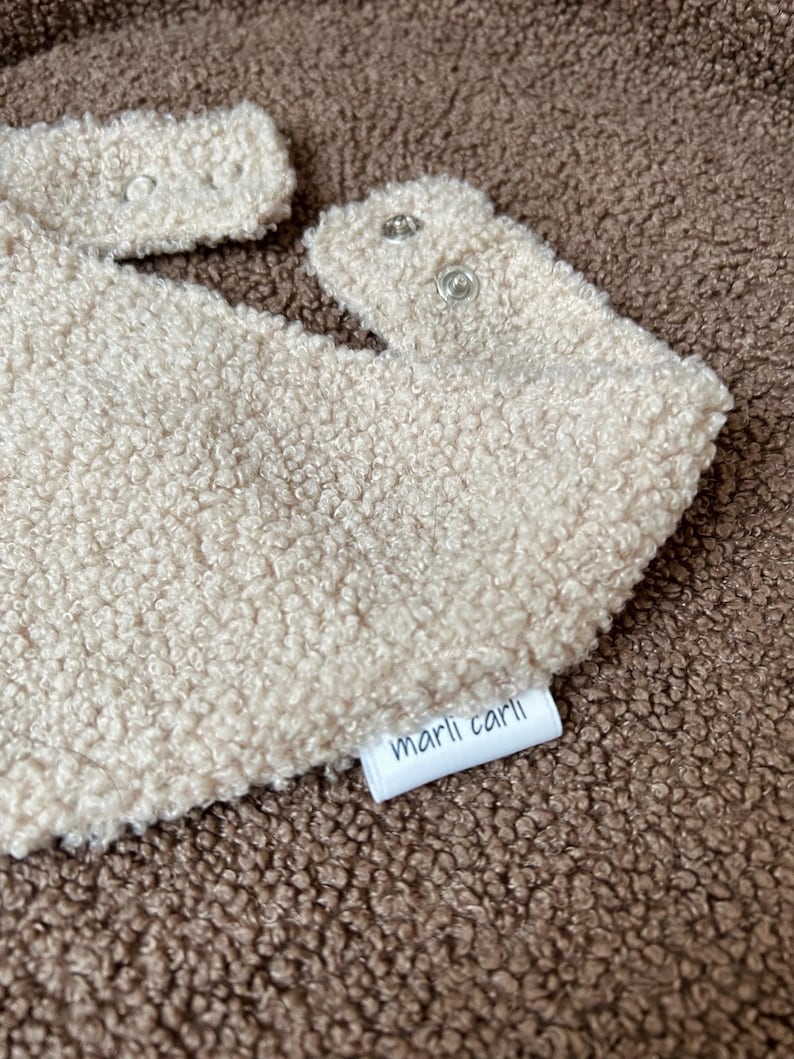 May include: A beige sherpa bib with a textured, fleece-like surface. The bib features two silver-toned metal snap closures at the neck and a small white fabric tag on the side with the text marli carli printed in black lowercase letters.