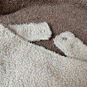 May include: A textured cream-colored fleece fabric featuring two silver-toned metal snap fasteners on a small tab, positioned against a background of dark brown textured fleece fabric. The material has a thick, curly, and soft appearance.