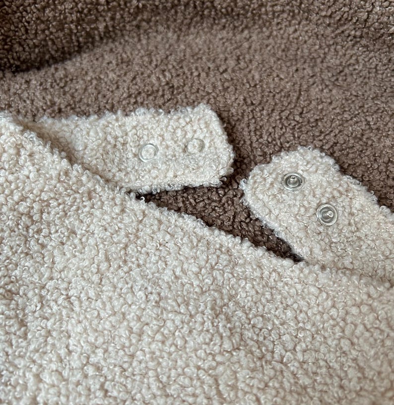 May include: A textured cream-colored fleece fabric featuring two silver-toned metal snap fasteners on a small tab, positioned against a background of dark brown textured fleece fabric. The material has a thick, curly, and soft appearance.