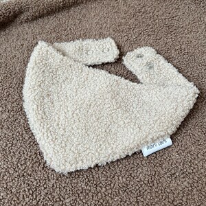 May include: A cream-colored bib made of textured, fuzzy sherpa fabric. The bib features a triangular shape with two snap closures at the top for fastening. A small white fabric tag with the black text marli carli is attached to the bottom edge.
