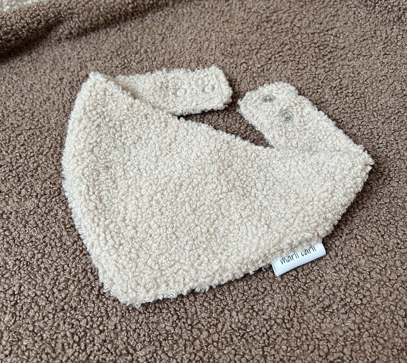 May include: A cream-colored bib made of textured, fuzzy sherpa fabric. The bib features a triangular shape with two snap closures at the top for fastening. A small white fabric tag with the black text marli carli is attached to the bottom edge.