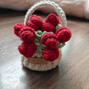 Könnte beinhalten: Ein handgefertigter Häkelkorb gefüllt mit roten Rosen und grünen Blättern. Der Korb ist weiß mit einem passenden Griff. Ein kleines braunes Etikett mit einem Bärenmuster ist am Korb befestigt.