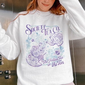 May include: White sweatshirt with a graphic design featuring the text "Society Tea Co." in purple script. The design includes a teapot, teacup, roses, and a bee, with the phrase "The Hottest Tea in the Ton."
