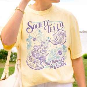May include: Pale yellow t-shirt with a floral and tea-themed graphic. The design includes a teapot, teacup, roses, and bees, with the text "Society Tea Co." and "The Hottest Tea in the Ton."
