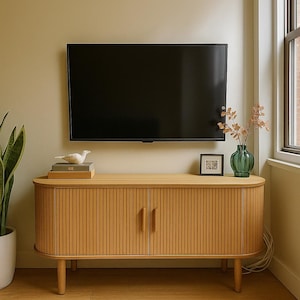 May include: A light wood TV stand with a ribbed front and two doors. A black flat-screen TV is mounted above the stand. Decorative items include a white bird figurine, books, a framed picture, and a green vase with dried flowers. A rug is on the floor.