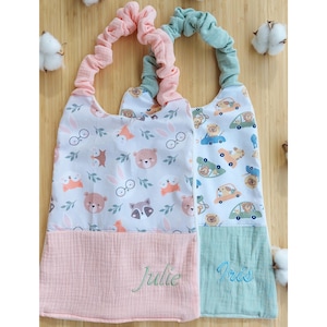 May include: Two baby bibs with fabric patterns and elastic neckbands. One bib has a pink neckband and a white pattern with bears, foxes, and glasses. The other has a green neckband and a pattern of cars and bears. The name "Julie" is embroidered on the pink bib.