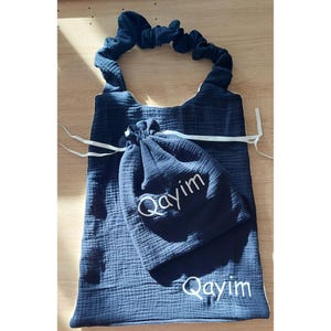 May include: A dark navy blue fabric bib with a gathered neck strap and a matching drawstring pouch. Both items feature the name Qayim embroidered in white thread. The fabric has a textured, crinkled appearance, and the pouch is tied with white ribbons.