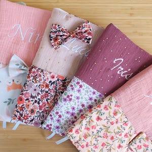 Peut inclure: Quatre housses en tissu rectangulaires avec des prénoms brodés, notamment Nina, Luce et Iris. Les housses sont composées de tissu mousseline uni et à motifs floraux, agrémentées de petits points dorés et de nœuds en tissu assortis.