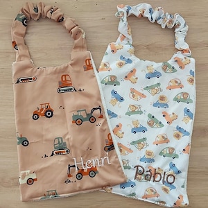 Puede incluir: Dos baberos rectangulares con aberturas elásticas para el cuello. El babero izquierdo es color canela con un patrón de vehículos de construcción y el nombre Henri bordado. El derecho es blanco con animales en coches y el nombre Pablo.