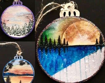 May include: Three round wooden ornaments with painted winter scenes. The first ornament has a purple glitter border and depicts a snowy forest with a light blue sky. The second ornament has a silver glitter border and depicts a forest with a green and blue sky and a large moon. The third ornament has a blue glitter border and depicts a sunset over a lake with mountains in the background.