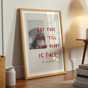 Puede incluir: Una impresión artística enmarcada con el texto "EAT THIS LIFE 'TILL YOUR HEART IS FULL" en rojo. La impresión tiene un marco de madera clara y está apoyada contra una pared blanca. Una imagen en blanco y negro está en el centro.