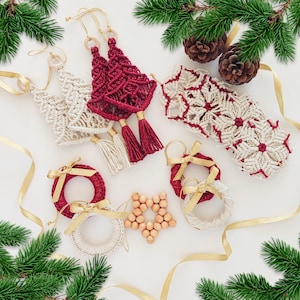 May include: A collection of handmade macrame ornaments including cream and deep red tree shapes with tassels, a cream and red snowflake-patterned runner, small circular wreaths with gold ribbons, and a star made of light wooden beads.