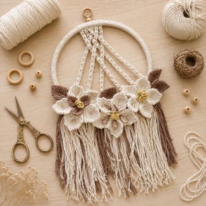 Può includere: Un arazzo in macramè con un cerchio rivestito in corda color crema, decorato con tre fiori complessi realizzati in filo color crema e marrone. Il design include motivi a corda annodata, accenti di foglie marroni e lunghe frange crema e marrone.