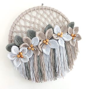 Puede incluir: Un tapiz de pared circular de macramé con un patrón de red beige tejido. La sección inferior está decorada con varias flores de tela en tonos gris, beige y blanco, acentuadas con pequeñas cuentas de madera y largos flecos multicolores.