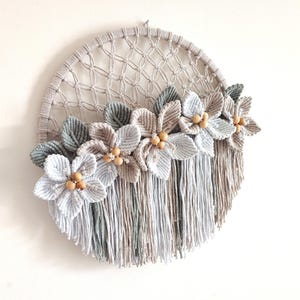 May include: A round macrame wall hanging featuring a woven net design in a light beige color. The wall decor is adorned with handmade flowers in shades of gray, beige, and green, with wooden bead centers and hanging fringe.