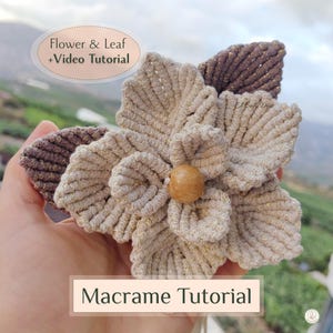 May include: A handmade macrame flower and leaf design in beige and brown tones. The flower has a wooden bead center and is surrounded by textured petals and leaves. The image includes the text "Flower & Leaf + Video Tutorial" and "Macrame Tutorial".