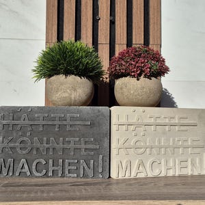 Könnte beinhalten: Zwei graue Beton Schilder mit dem Text "HATTE KONNTE MACHEN!" darauf. Die Schilder stehen auf einem Holztisch.