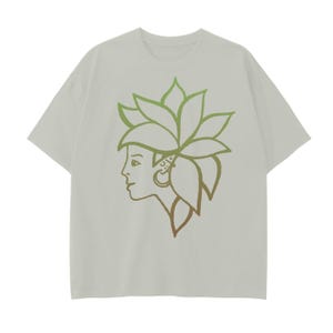 以下が含まれることがあります： 薄いグレーのTシャツに、緑と茶色の輪郭で描かれた女性の顔と、頭に葉っぱのデザインが描かれている