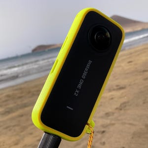 May include: A black Insta360 ONE X2 camera with a bright yellow protective case is mounted on a black selfie stick. The camera is held at an angle, with the ocean and beach in the background. A yellow and black lanyard is attached to the camera.