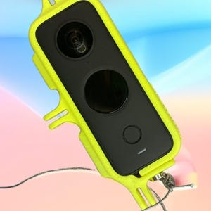 Puede incluir: Una cámara de acción negra con una funda protectora de color amarillo brillante. La cámara tiene una lente, un botón negro grande y un botón más pequeño. Un fino cordón gris está sujeto a la funda.
