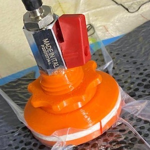 Könnte beinhalten: Nahaufnahme eines orangefarbenen Kunststoffventils mit einem silbernen Metallanschluss und einem roten Hebel. Das Ventil ist mit "MADE IN ITAL P200707934L" beschriftet.