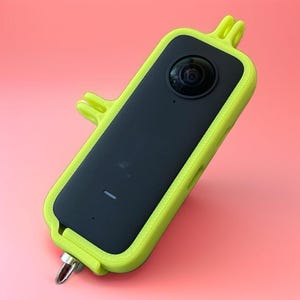 Könnte beinhalten: Eine schwarze und hellgelbe Action-Kamera mit Schutzhülle. Die Kamera hat ein einzelnes Objektiv und einen kleinen Knopf. Die Hülle ist hellgelb und hat eine Schlaufe zum Anbringen eines Lanyards. Der Hintergrund ist einfarbig rosa.