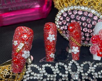 May include: Five red and white glitter nail designs with rhinestones and small flowers. The nails are long and have a pointed shape.